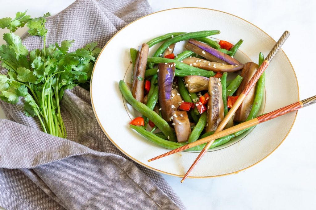 Eggplant, green beans, and red peppers in a white bowl with chopsticks. Garnished with sesame seeds and a bunch of cilantro on a grey napkin.