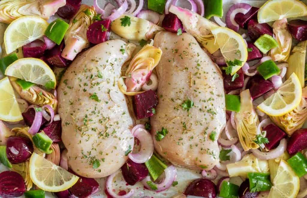 Chicken with beets, artichokes, peppers, onion, and lemons on a sheet pan with seasonings.