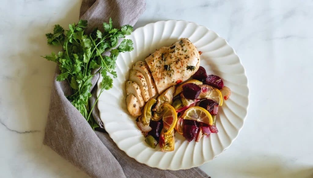 Chicken with beets, lemons, onions, and artichokes on a white plate with parsley and a grey napkin.